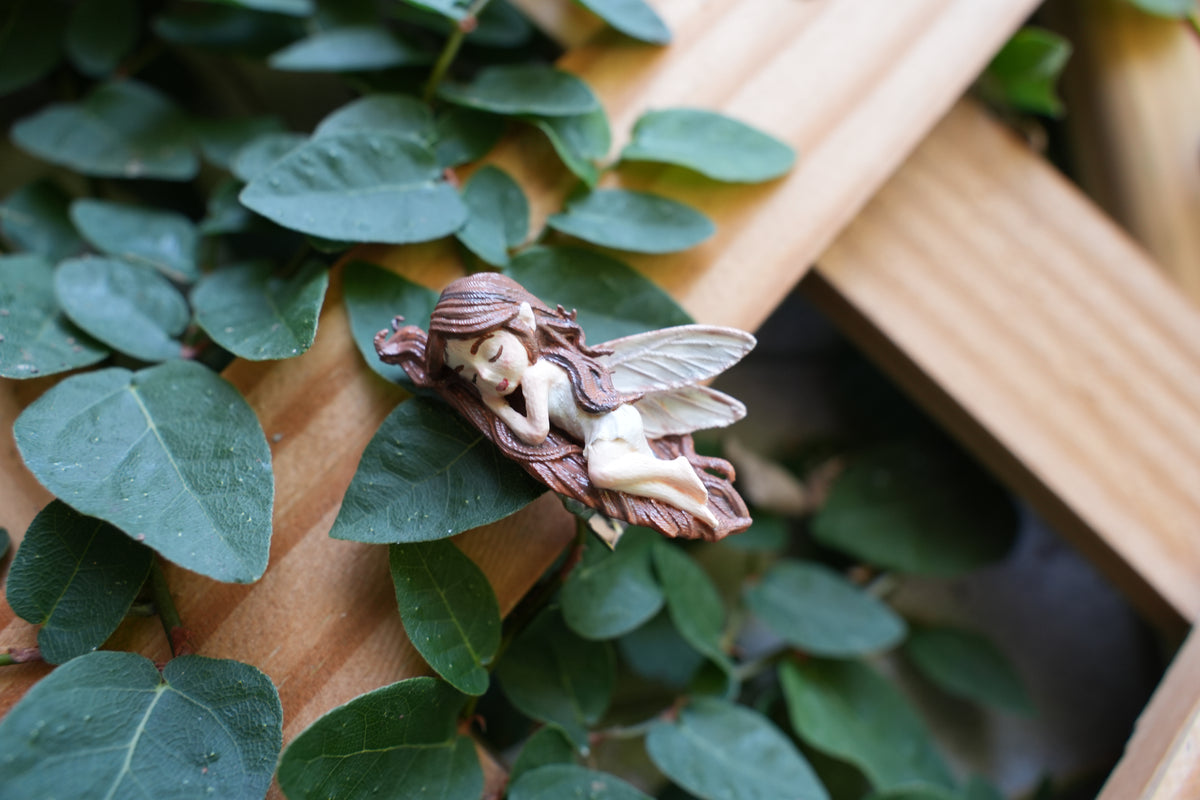 A small fairy garden figurine of a sleeping female fairy with brown hair and wings, resting on a wooden trellis surrounded by green ivy leaves.